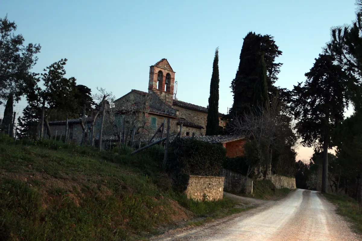 Chiesa in pietra con campanile a vela tra cipressi, vista da una strada di campagna